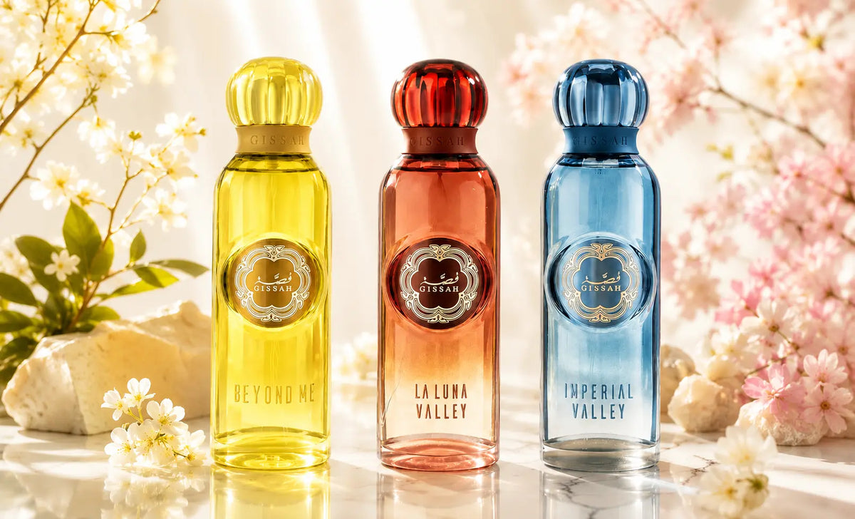 Three tall perfume bottles from Royal Oud are lined up on a reflective surface with soft white and pink blossoms. The left bottle is yellow and labelled 'BEYOND ME', the middle is orange-red and labelled 'LA LUNA VALLEY', and the right is blue and labelled 'IMPERIAL VALLEY'.
